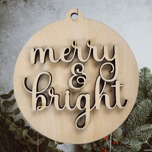 A layered "merry and bright" wood craft kit shown assembled on a bed of holiday greenery.