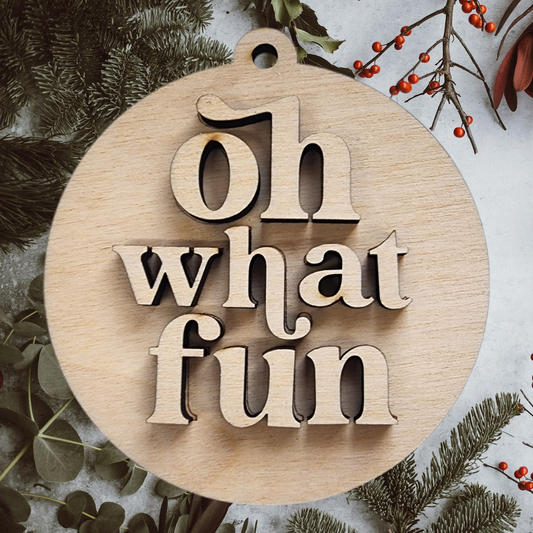 A layered "oh what fun" wood craft kit shown assembled on a bed of Christmas greenery.